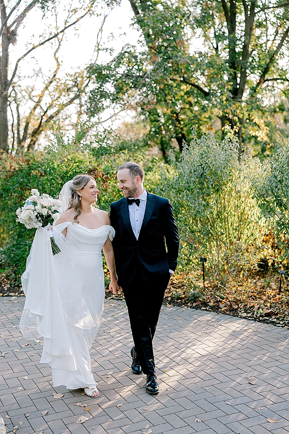 Couple portrait of bride and groom walking hand in hand on a brick garden path, bride gazing at groom with veil and bouquet