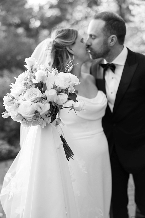 Wedding kiss in a black and white wedding photo, bride in veil holding bouquet as groom in tuxedo kisses her amid soft greenery bokeh