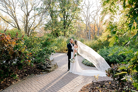 Couple portrait of bride and groom embrace on a sunlit garden brick path, her long cathedral veil blowing behind her bouquet