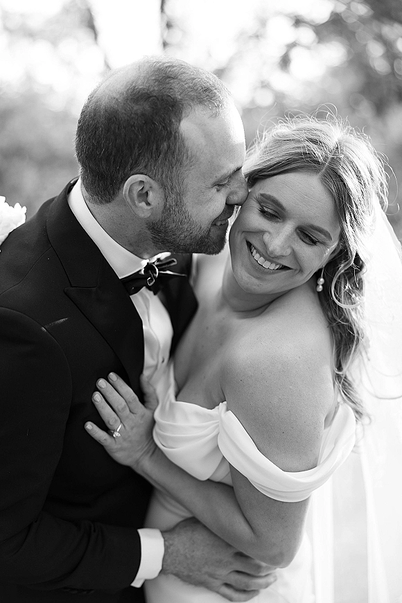 Wedding couple portrait of groom kissing bride’s cheek as she smiles, veil and strapless dress glowing in sunlit trees backdrop