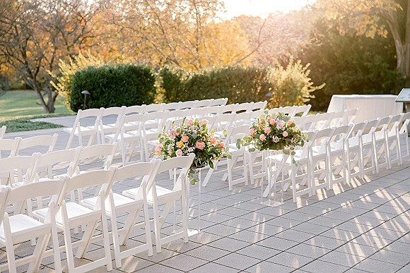 Ceremony seating with outdoor wedding ceremony chairs lined in rows, rose and eucalyptus florals on clear stands along a sunny stone patio aisle