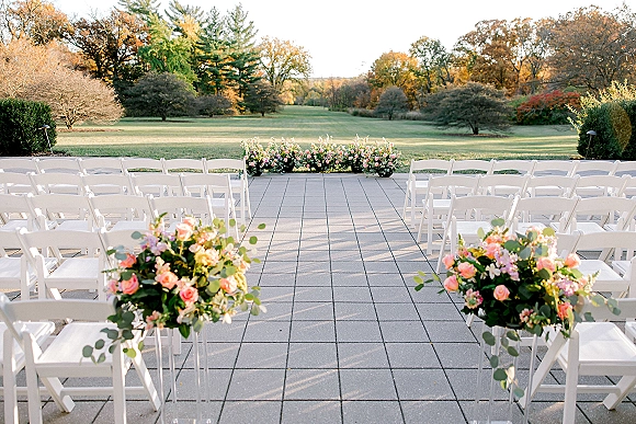Ceremony aisle decor with white folding chairs and pink rose florals lining a wide aisle on a stone patio beside autumn trees