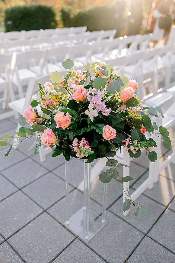 Ceremony aisle flowers in a pastel arrangement of pink roses and eucalyptus on an acrylic pedestal beside white folding chairs outdoors
