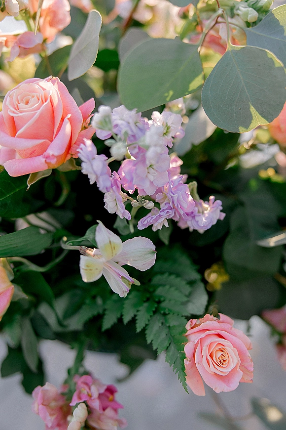 Wedding floral arrangement with a pink rose centerpiece of lavender and white blooms, eucalyptus, and fern fronds against sunlit greenery