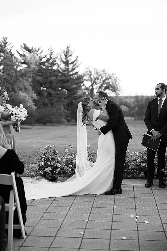 Wedding kiss portrait of bride and groom kissing, her long veil and off-the-shoulder gown framed by florals on an outdoor lawn ceremony aisle