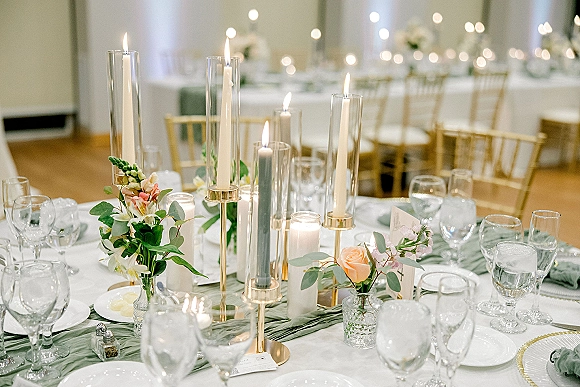 Reception tablescape with taper candle centerpiece, greenery runner, and small florals on white linen, set in a banquet hall with string lights