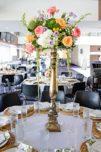 Tall wedding centerpiece in a gold pedestal vase showcasing elevated centerpiece ideas amid round tables in a reception hall with windows.