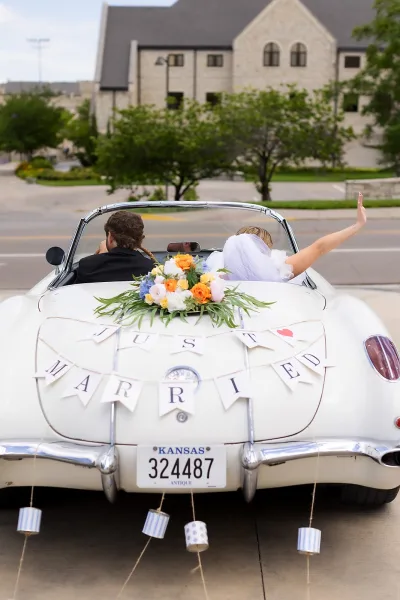 Getaway car with a just married sign, tin cans trailing as the couple drives off, passing a stone building and trees on the quiet street.