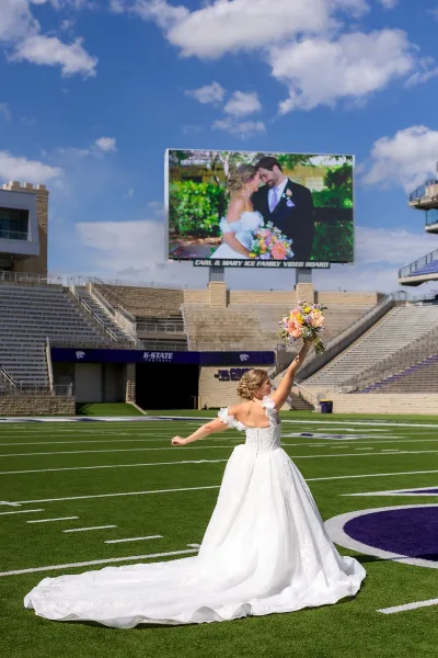 Bridal portrait in a football field wedding photo, the bride’s dress train sweeps the turf beneath stadium seating and a sunlit blue sky.