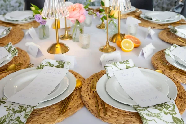 Reception tablescape featuring an elegant wedding place setting with rattan chargers on a white tablecloth, set before floral wallpaper.