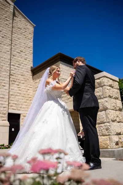 Bride pinning boutonniere on her groom in a candid wedding moment, her long lace veil flowing outside a stone building under a blue sky.