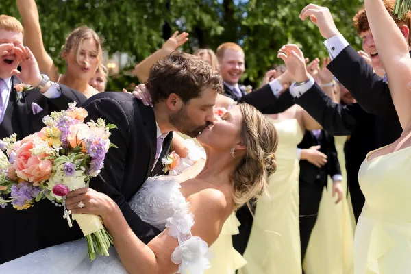 Wedding kiss portrait as the groom dips the bride, bouquet aloft, surrounded by a cheering wedding party on a sunlit lawn under trees.