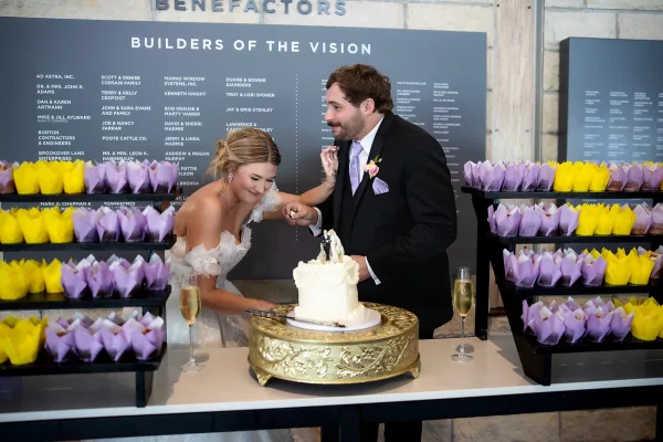 Wedding cake cutting as a couple feed each other cake beside a small single-tier cake on a gold cake stand by a donor wall inside.