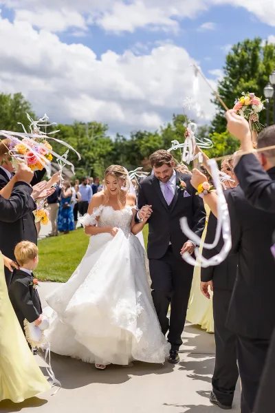 Wedding exit through a ribbon wand exit as the couple hold hands, guests cheer along a garden pathway beneath blue sky and trees.