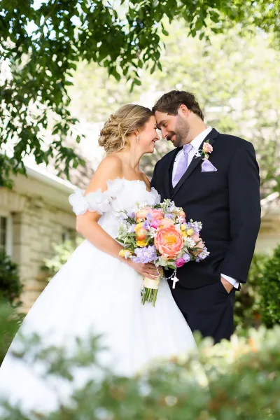 Couple portrait of a bride and groom sharing a forehead touch as she holds a pastel bouquet amid garden trees by a stone building.