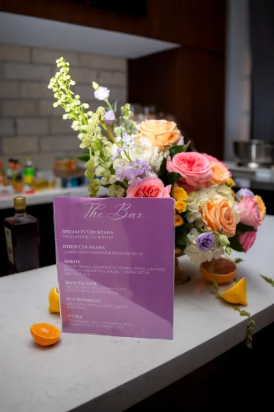 Wedding bar sign with an acrylic bar menu, styled with fresh citrus, on a bar counter against a tiled wall with liquor bottles and glassware.