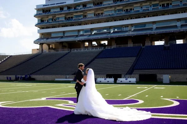 Couple portrait of a bride and groom embracing on a football field, her veil trailing as stadium bleachers rise beneath a bright open sky.
