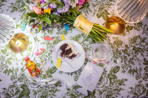 Reception table details with a wedding cake slice on a plate and a bridal bouquet atop a floral tablecloth.