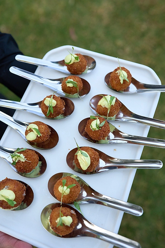 Wedding passed appetizers on a white tray, cocktail hour appetizers served on tasting spoons with fried bites, creamy sauce, and microgreens on a lawn