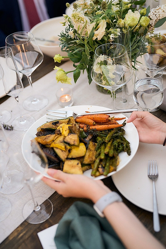 Wedding place setting with white plates, linen napkins, wine glasses, and a floral greenery centerpiece on a wood table with votive candle