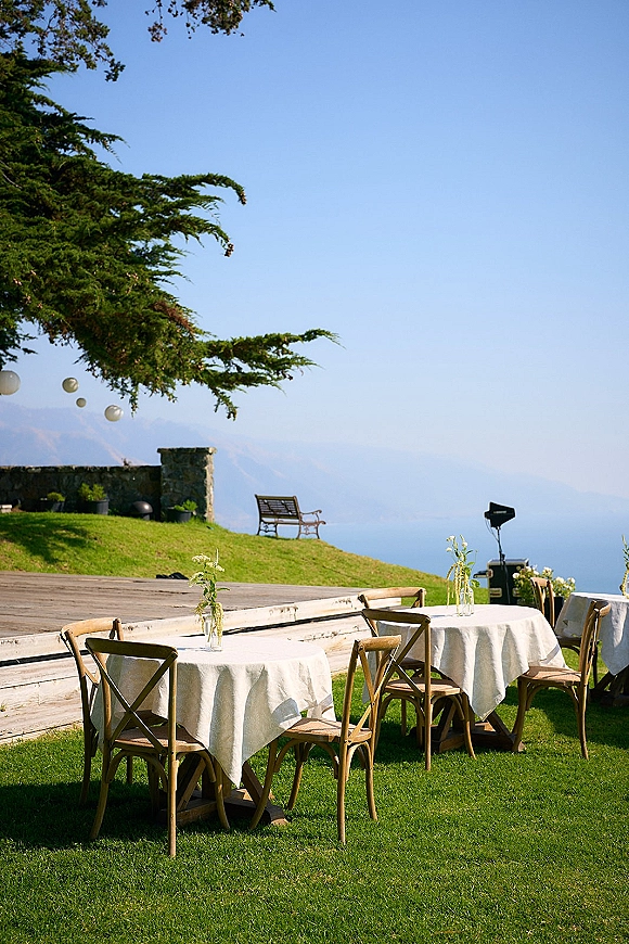 Outdoor reception tables with white linens and wooden cross back chairs, bud vases and greenery on a lawn with lanterns and mountains