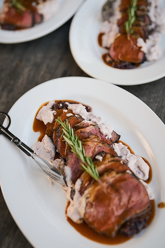 Wedding dinner plate with sliced beef, brown and cream sauce, and rosemary garnish on a white plate on a dark wood table