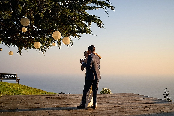 First dance outdoors as bride in a strapless wedding dress and groom in a dark suit sway under paper lanterns by the ocean at dusk
