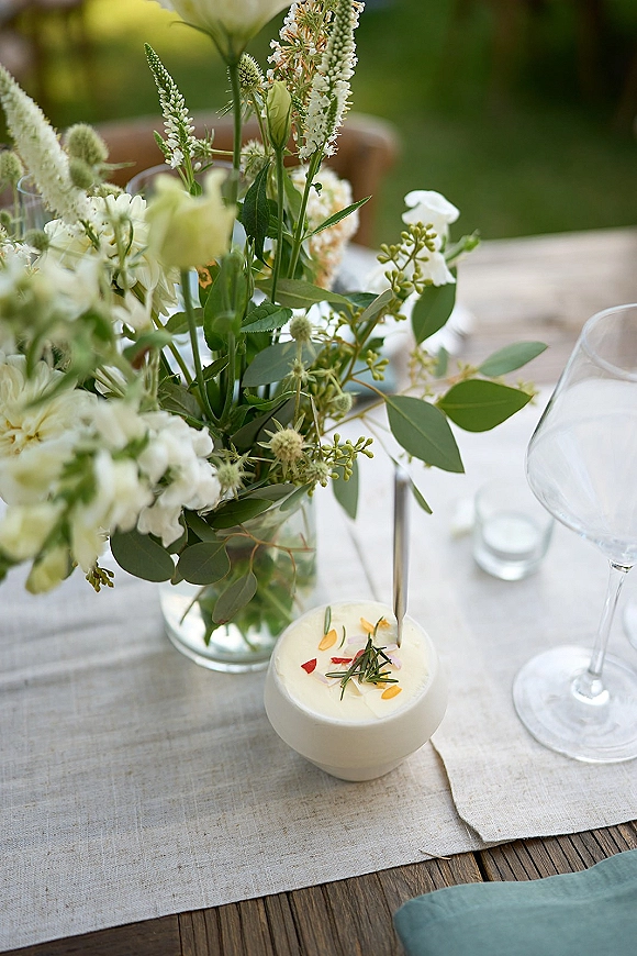 Reception tablescape with a wedding table centerpiece of white flowers and greenery in a glass vase, linen runner, wine glasses, and a taper candle outdoors
