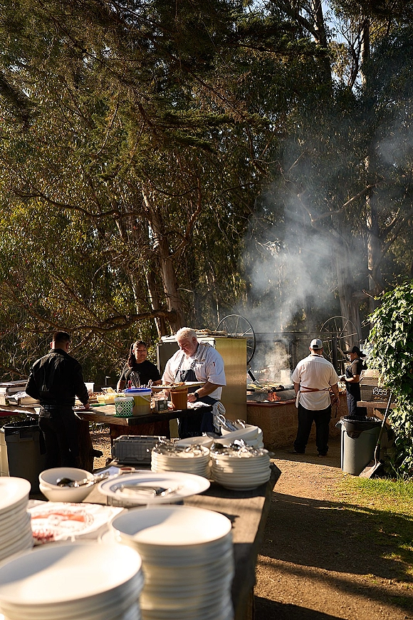 Wedding catering staff in chef coats preparing food at an outdoor wedding catering grill station with smoke in a sunlit wooded setting