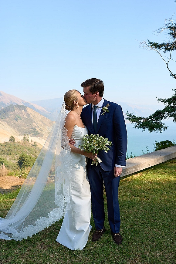 Wedding kiss portrait of bride and groom kissing outdoors, her lace veil blowing as she holds a bouquet with mountain and ocean views behind