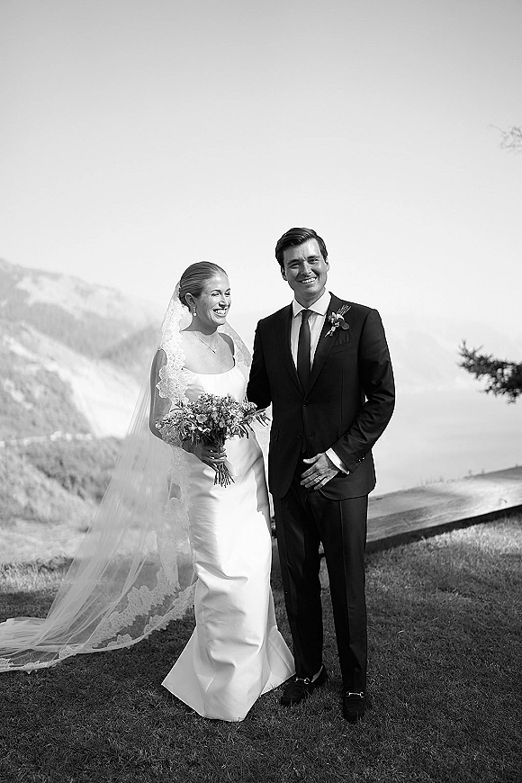 Couple portrait in a black and white wedding portrait style, bride with lace veil and bouquet beside groom in suit by a mountain lake