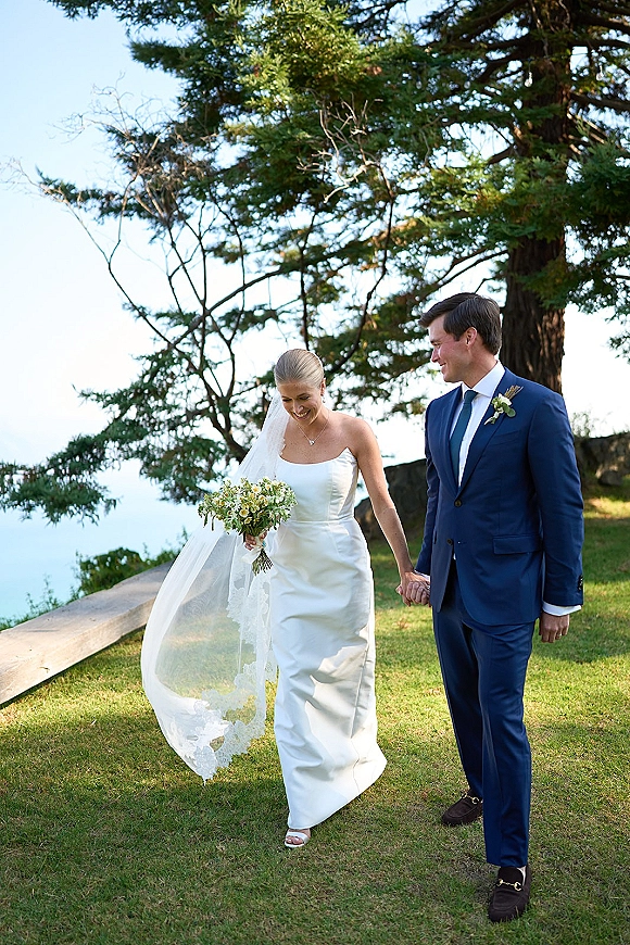 Couple portrait of bride and groom holding hands, her veil blowing as they walk on a grassy lawn by water and trees