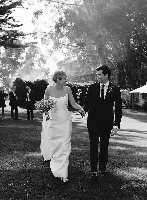 Newlywed couple walking hand in hand, bride holding bouquet, groom in suit, on a sunlit lawn walkway with guests behind them