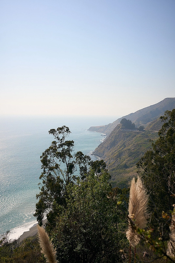 Coastal cliff view with an ocean cliff overlook showing rugged rocky coastline, green hills, trees, and blue horizon under clear sky