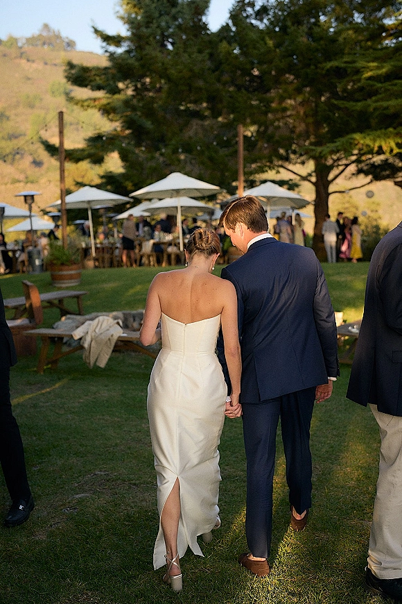 Wedding couple walking away hand in hand, bride in strapless gown and groom in navy suit on a lawn reception with white umbrellas