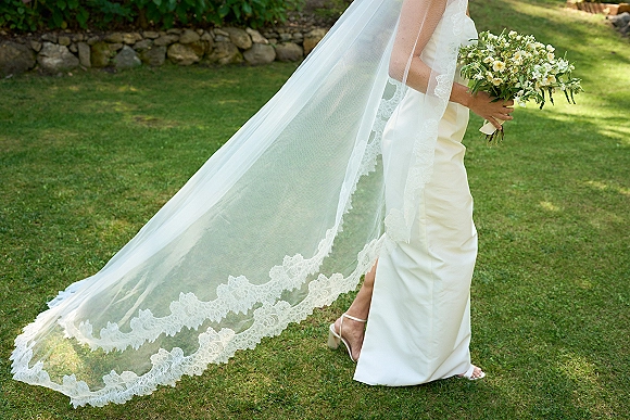 Bridal portrait with long wedding veil trailing behind, bride in strapless gown holding white and green bouquet on a garden lawn by stone wall