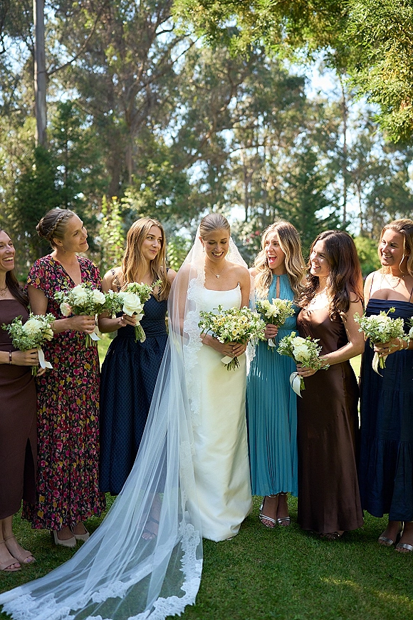 Bride with bridesmaids laughing in a bridal party portrait, holding white and green bouquets on a sunny lawn with garden trees behind