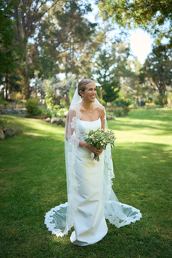 Bridal portrait of a smiling bride holding bouquet in a strapless dress and lace veil on a daylight garden lawn with trees