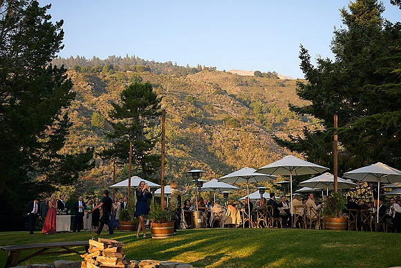 Outdoor wedding reception with backyard wedding reception seating under string lights, patio umbrellas and heaters on a lawn with mountain hills and pines