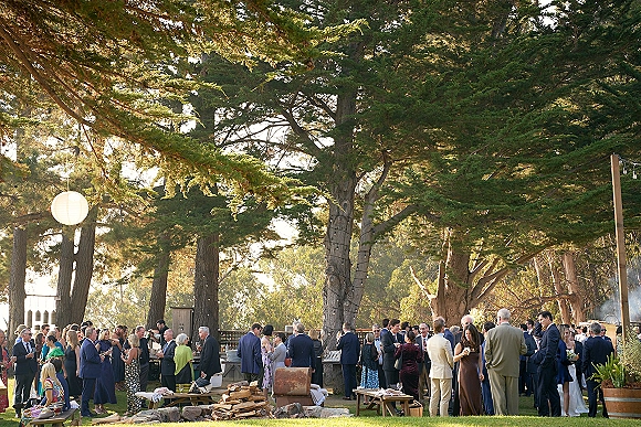 Wedding cocktail hour with guests mingling around an outdoor cocktail hour bar, wooden benches and picnic tables under string lights on a sunny lawn