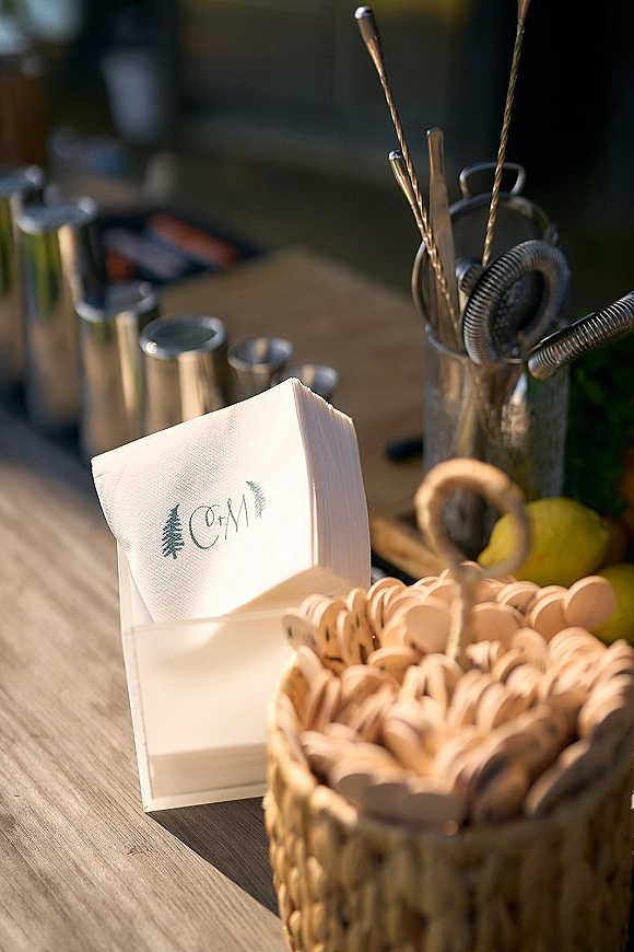 Wedding cocktail bar with signature cocktail napkins, shaker and jiggers neatly arranged on a wood counter with blurred bar setup behind