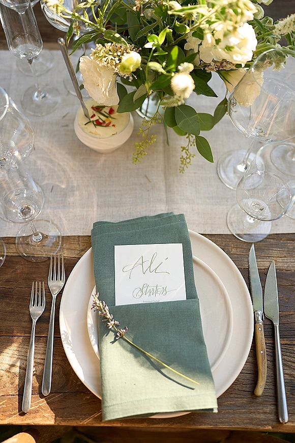 Reception tablescape with wedding place setting, green linen napkin, white floral centerpiece, taper candle, and glassware on a wooden table runner