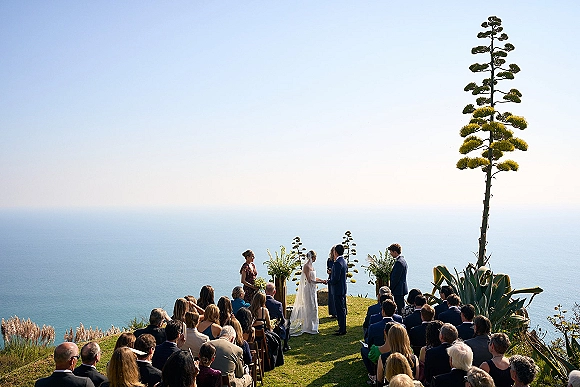 Wedding ceremony with bride in veil and groom in suit facing officiant, guests on wooden chairs on a cliffside lawn with ocean backdrop