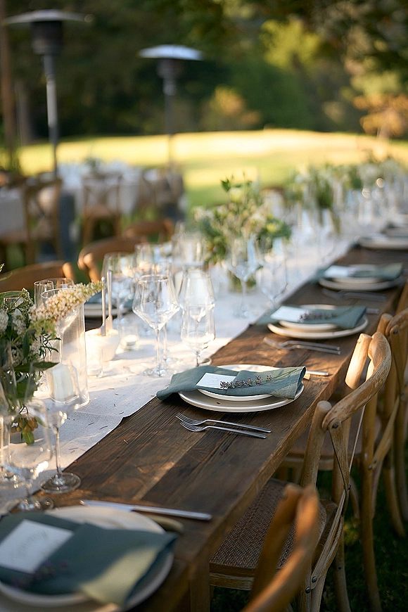 Reception tablescape with an outdoor wedding reception table set on a wood farm table, white runner, lavender sprigs, and greenery vases on a sunlit lawn