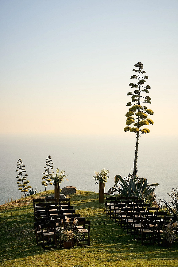 Ceremony setup with outdoor ceremony seating in black folding chairs, wood plinth florals and aisle flowers on a grassy oceanfront cliff lawn