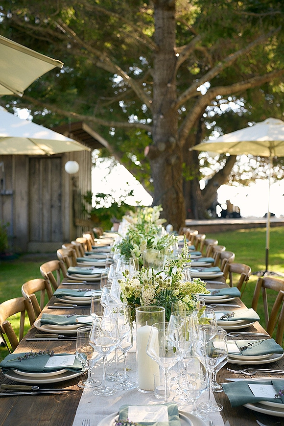 Reception tablescape with long banquet table, white runner, sage napkins, floral greenery, candles, and string lights by a rustic building