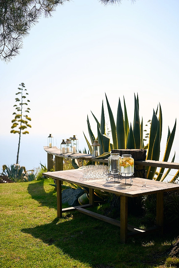 Wedding drink station on a wood table with glass beverage dispensers, citrus slices, and lantern candles overlooking the ocean lawn