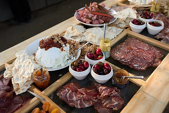 Charcuterie board with wedding cocktail hour food like sliced salami, cheeses, olives, crackers, and chutney jars on a wooden board tabletop