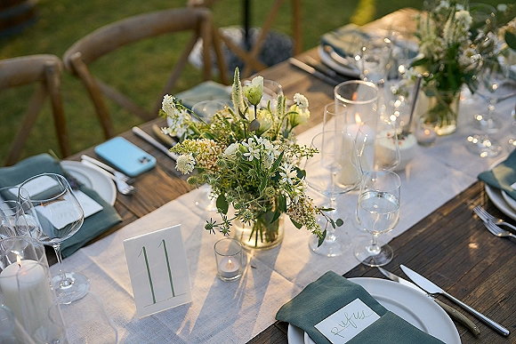 Reception tablescape on a rustic wedding table with linen runner, green and white florals, bud vases, candles, and string lights on lawn grass