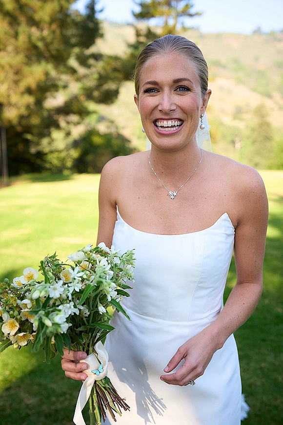 Bridal portrait of a smiling bride in a strapless wedding dress with veil, holding a white-and-green bouquet on a lawn with hills beyond